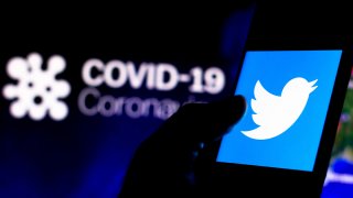 The Twitter logo is displayed on a smartphone with a computer model of the COVID-19 coronavirus in the background.