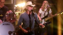 Kenny Chesney Performing on The Tonight Show with a guitar in his hand. A guitarist and drummer are behind him.