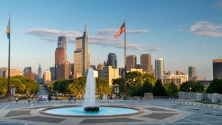 A fountain shoots water into the air as a pole with the American flag, as well as a row of skyscrapers, can be seen in the background of the Philadelphia skyline.