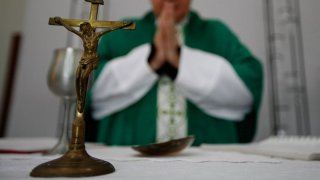 A crucifix sits on a table in front of a priest who is wearing a robe and has his hands clasped in prayer.