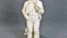 A carved, white marble sculpture of a boy holding a parrot. He leans against a pillar.