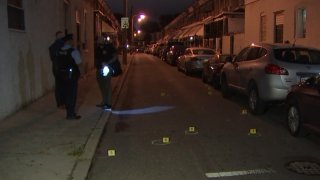 Philadelphia Police Department investigators stand on a sidewalk as bullet casings and evidence markers lay on the ground after a shooting.