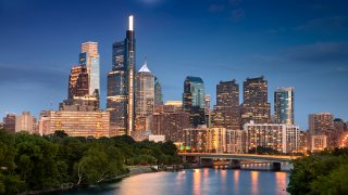 Various skyscrapers are shown in front of a body of water as part of the Philadelphia skyline at dusk.