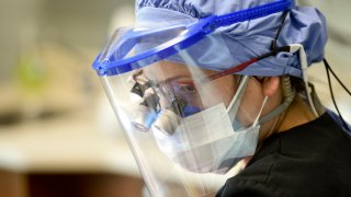 A dentist wears a face shield over a face mask as she looks on at a patient.