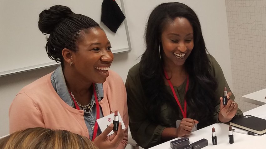 Amanda Johnson (left) and KJ Miller (right) smile as they sit at a table and look on at lipsticks.