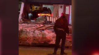 A man looks on in the distance, where a car can be seen having rammed through a house.