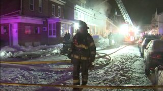 A firefighter walks along a Trenton, New Jersey, street. Behind him can be seen row houses on the left and parked cars on the right. In the middle of the street is a fire truck with its ladder expanded upward as firefighters douse a home with water from above.