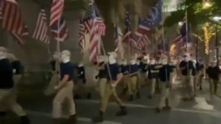 A group of men wearing kakis, blue shirts and white face coverings march next to Philadelphia City Hall while waving flags derivative of the American flag. The group is part of the white supremacist Patriot Front organization.