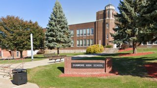 "Dunmore High School" is written on a small wall that's placed on a patch of grass. Trees surround the wall and behind the tree rises a multi-story building.