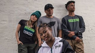 A woman in a zip-up jacket with the letters "PHI" embroidered on it crouches as another woman and two men stand behind her. The woman on the far left wears a t-shirt saying "Go Birds," as well as a beanie. The man in the center wears a gray t-shirt with a black logo of the Philadelphia Eagles. The man on the right wears a sweater with the words "go birds" printed across the chest.