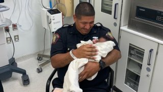 A police officer cradles a baby wrapped in a blanket while sitting in a hospital room.