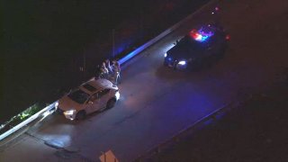 A white sedan pulls over to the side of the road as a police cruiser parks behind it with its lights on. Officers speak to the driver, who sits on a guardrail.