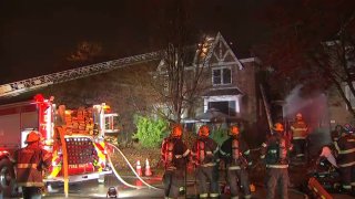 Firefighters and a fire truck stand in front of a two-story house. The truck's ladder extends toward the roof.