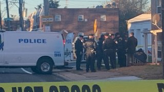 Police officers and homicide investigators look down the stairwell of an apartment where two men were found dead. A police van is parked next to the officers. An apartment building can be seen in the background.