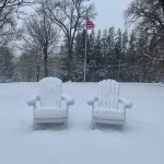 Snow-covered lawn chairs as an American flag on a pole waves behind.