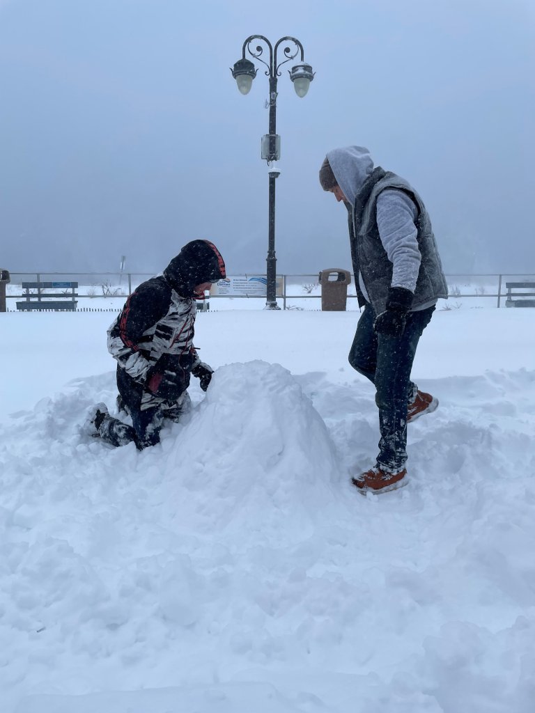 A man on the left kneels next to a pile of snow while a man stands on the other side of the pile.