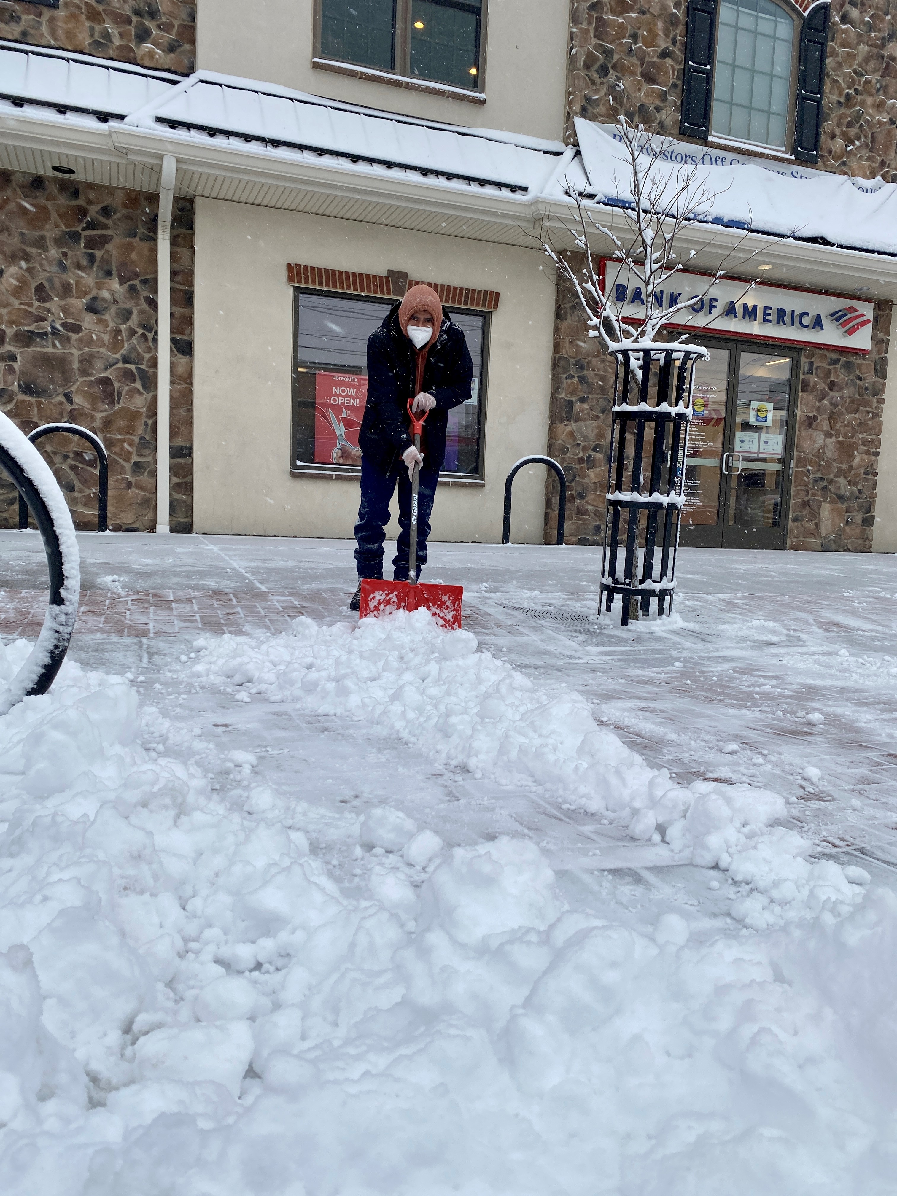 A man in a hoodie and wearing a face mask shovels snow from in front of a building in Delaware.