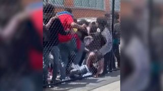 A group of teens is seen beating a grounded teen who is up against a chain-link fence.