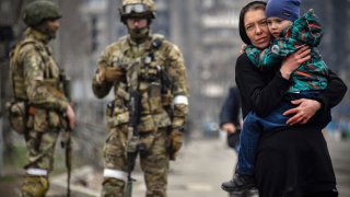 A woman holds a child next to Russian soldiers in a street of Mariupol on April 12, 2022, as Russian troops intensify a campaign to take the strategic port city, part of an anticipated massive onslaught across eastern Ukraine, while Russia’s President makes a defiant case for the war on Russia’s neighbour. – *EDITOR’S NOTE: This picture was taken during a trip organized by the Russian military.*