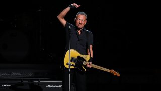 Bruce Springsteen wearing guitar and pointing finger in sky.