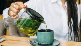 A woman pours green tea from a glass teapot.