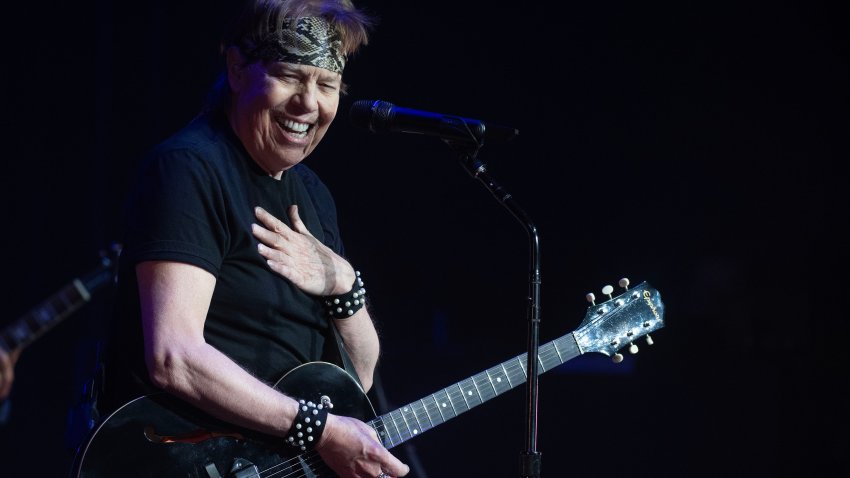 George Thorogood with guitar on holds hand over chest while smiling.