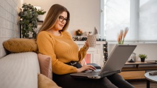 Young smiling woman sitting on a couch, working on her laptop and holding a cup of coffee