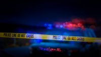 A stock photo of a Police Line "Do Not Cross" caution tape with a defocused police car with sirens flashing red and blue.