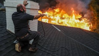 Altadena, CA, Wednesday, Jan 8, 2025 – Jimmy Orlandini shields from intense heat as he hoses down a neighbors rooftop on Sinaloa Ave. as the Eaton Fire continues to grow. 