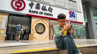 SHENZHEN, CHINA – NOVEMBER 16: A boy sits outside a branch of the Bank of China while using a smartphone on November 16, 2024 in Shenzhen, Guangdong Province, China.  