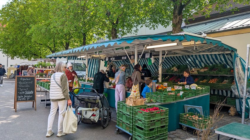 Shoppers buy fresh vegetables, fruit, and herbs at an outdoor produce market under green-striped canopies in Regensburg, Upper Palatinate, Bavaria, Germany, on April 19, 2025.